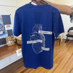 Blue T-shirt on a hanger featuring a white abstract heart design and Ukrainian words Людяність, Професійність, Довіра.
