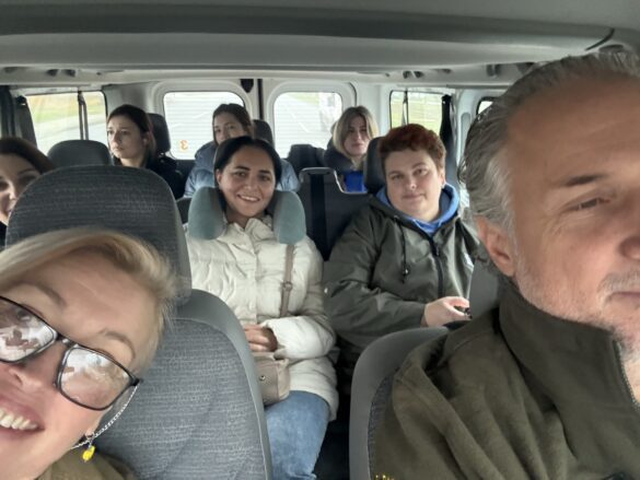 Smiling group of eight adults seated in a van, taking a casual selfie from the front passenger area.
