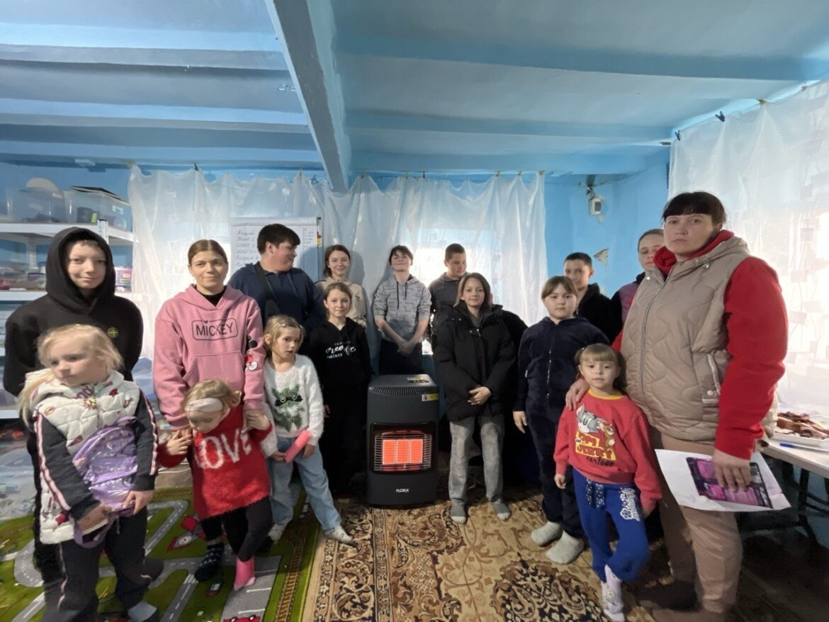 Group of children and adults posing together around a small heater in a blue room with plastic curtains behind them, some wearing jackets and hoodies.